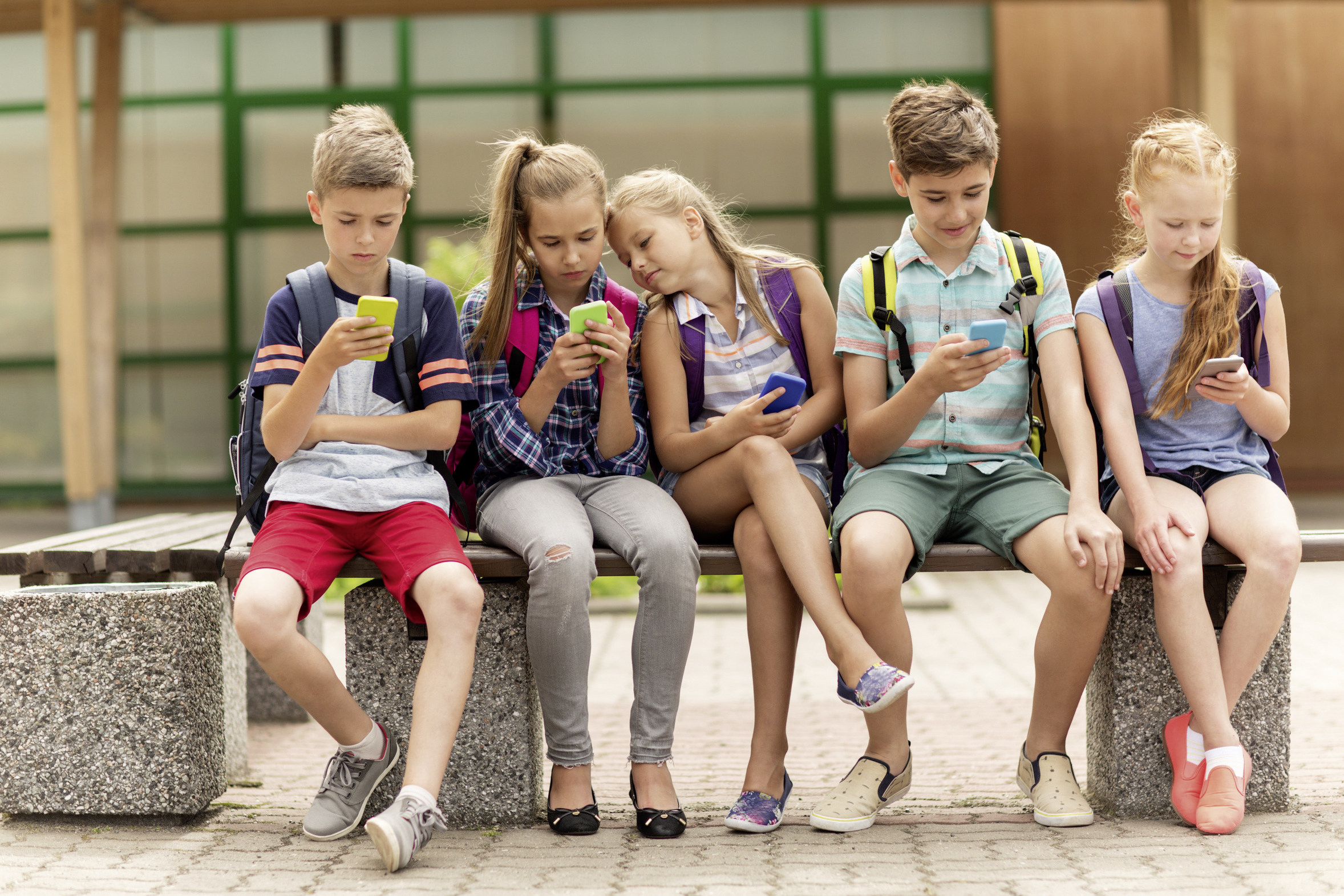elementary school students with smartphones / lev dolgachov primary education, friendship, childhood, technology and people concept - group of happy elementary school students with smartphones and backpacks sitting on bench outdoors